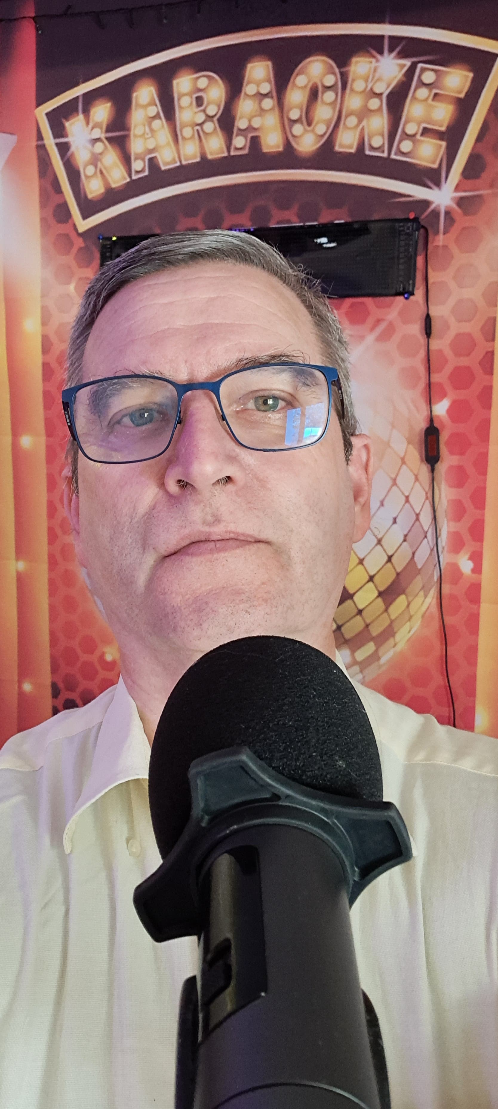 Homme à lunettes devant un micro, fond avec enseigne karaoké lumineuse et boule disco.
